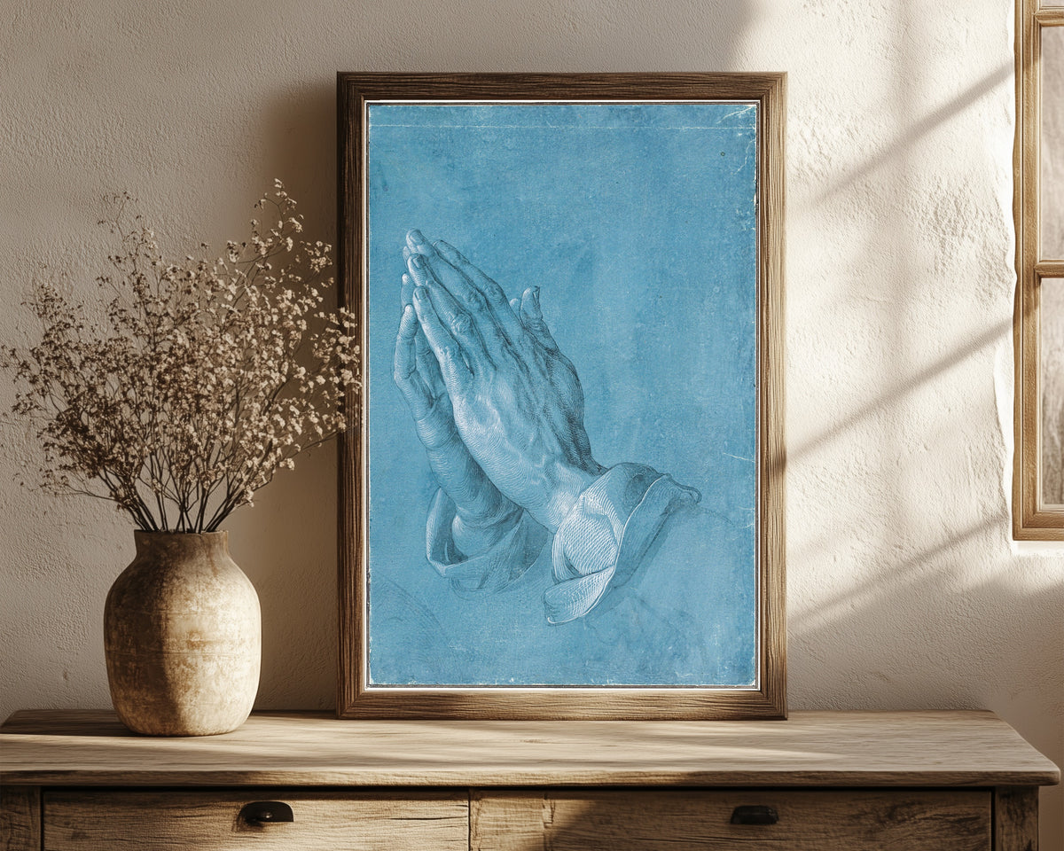 Framed art print of Praying Hands by Albrecht Dürer, in a dark wood frame leaning on a rustic console beside dried flowers