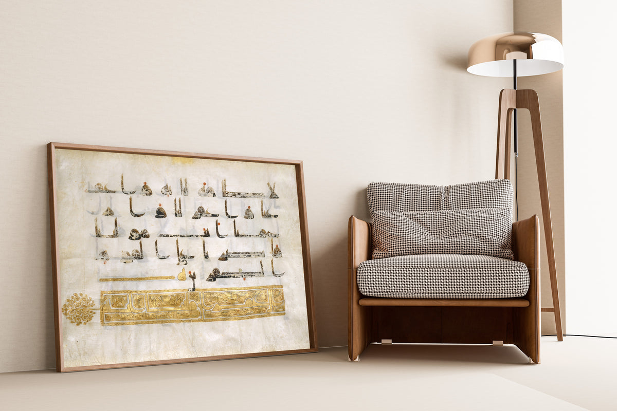 Framed art print of Qur'an Folio (Gold Heading Band), leaning against a wall beside an armchair and a floor lamp