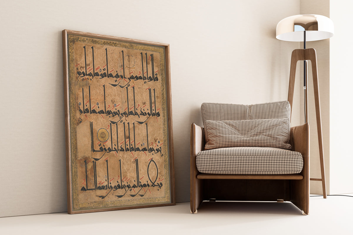 Framed art print of Qur'an Folio (Gold Roundel on Arabesque Ground), leaning against a wall beside an armchair and a floor lamp