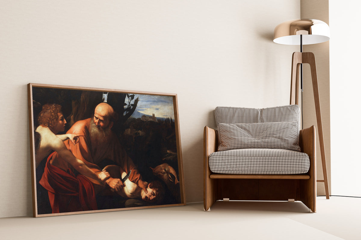 Framed art print of Sacrifice of Isaac by Caravaggio, leaning against a wall beside an armchair and a floor lamp