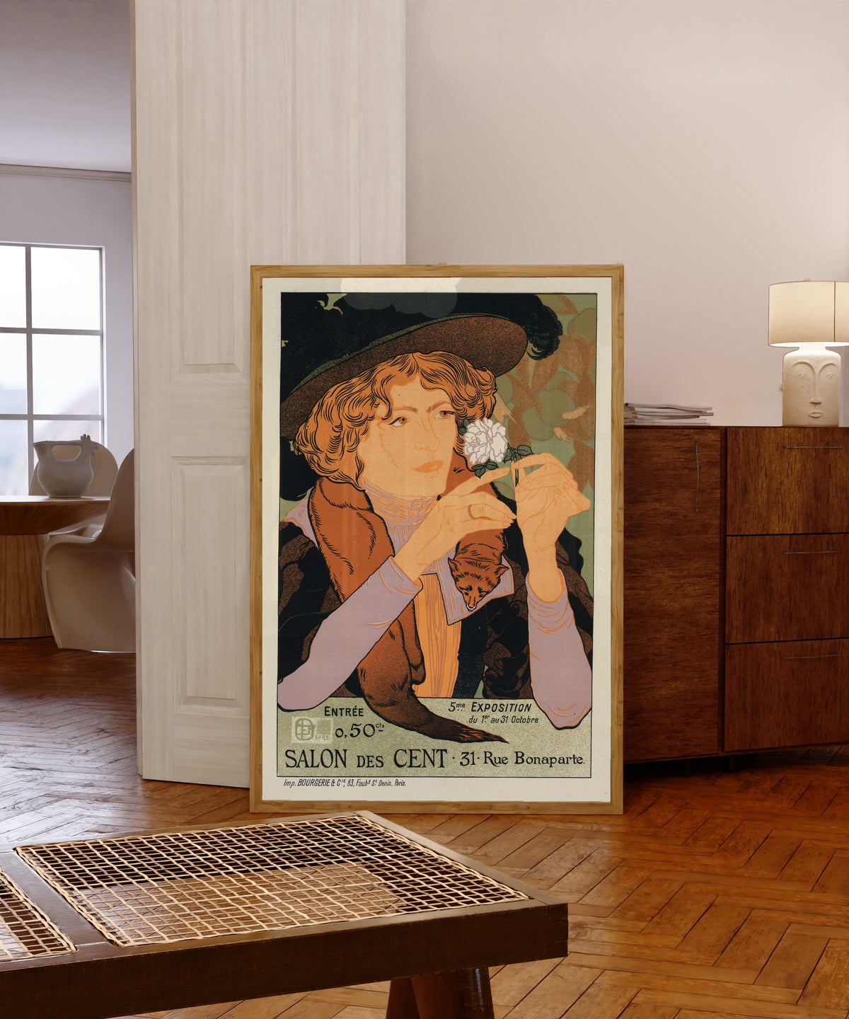 Framed art print of Salon des Cent: 5e Exposition d'Art by Georges de Feure, in a light wood frame leaning in a warm room with a parquet floor