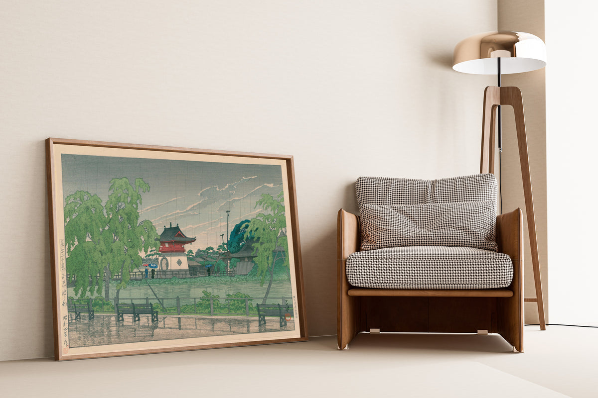 Framed art print of Shinobazu Pond in Rain by Kawase Hasui, leaning against a wall beside an armchair and a floor lamp