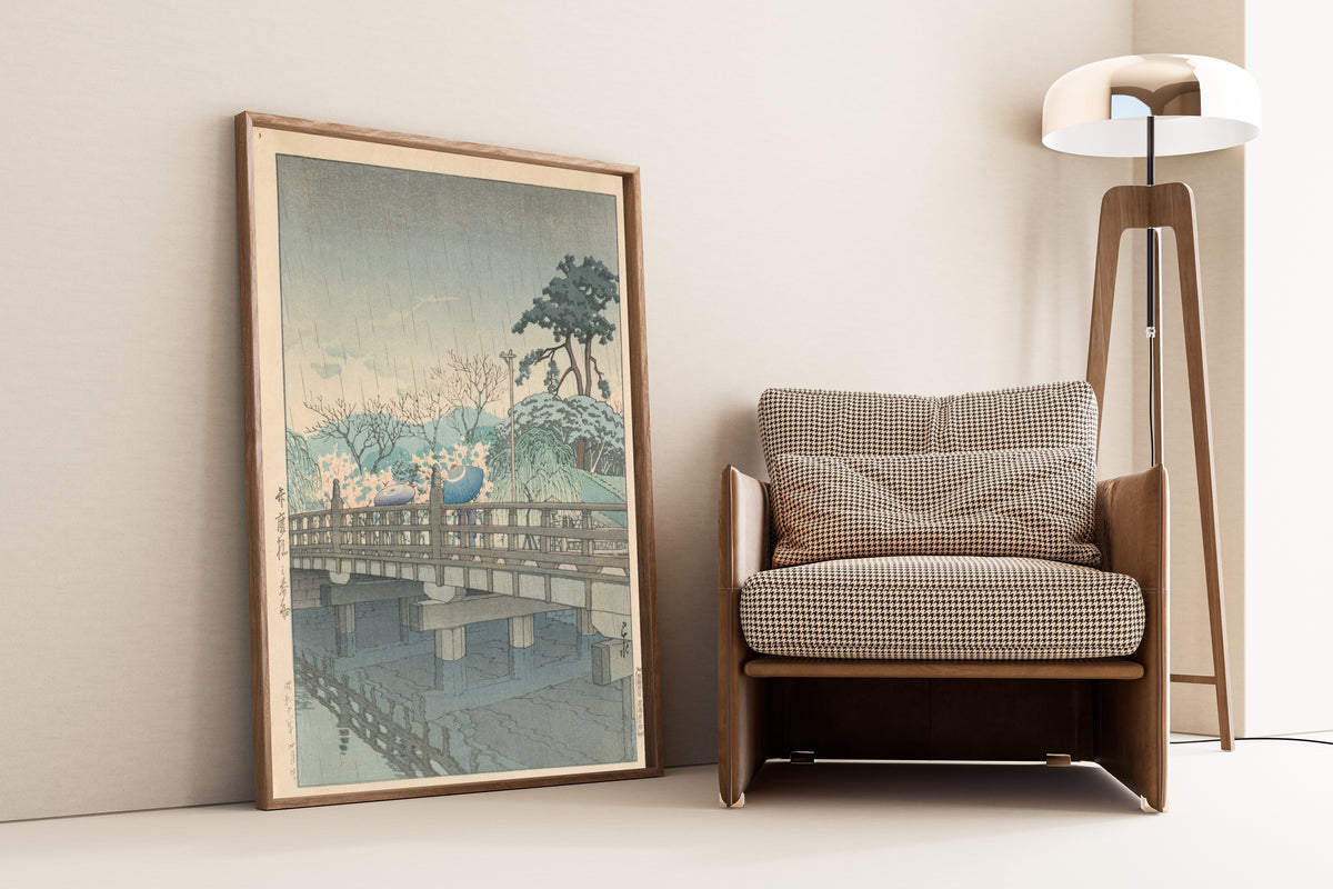 Framed art print of Spring Rain at Benkei Bridge by Kawase Hasui, leaning against a wall beside an armchair and a floor lamp