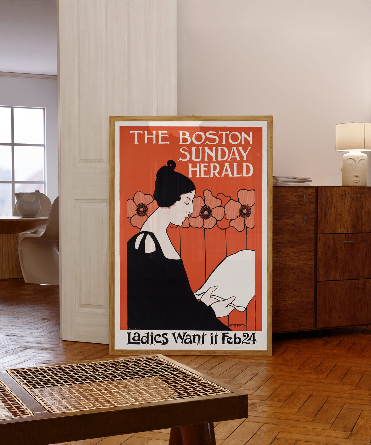 Framed art print of The Boston Sunday Herald, Ladies Want It, Feb 24 by Ethel Reed, in a light wood frame leaning in a warm room with a parquet floor