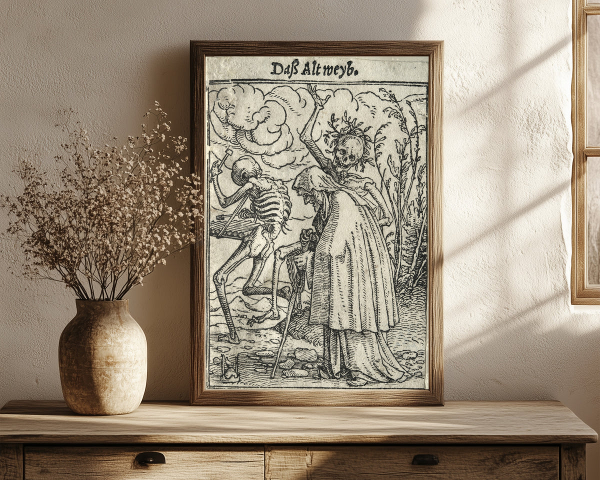 Framed art print of The Dance of Death: Daß Altweyb (The… by Hans Holbein the Younger, in a dark wood frame leaning on a rustic console beside dried flowers