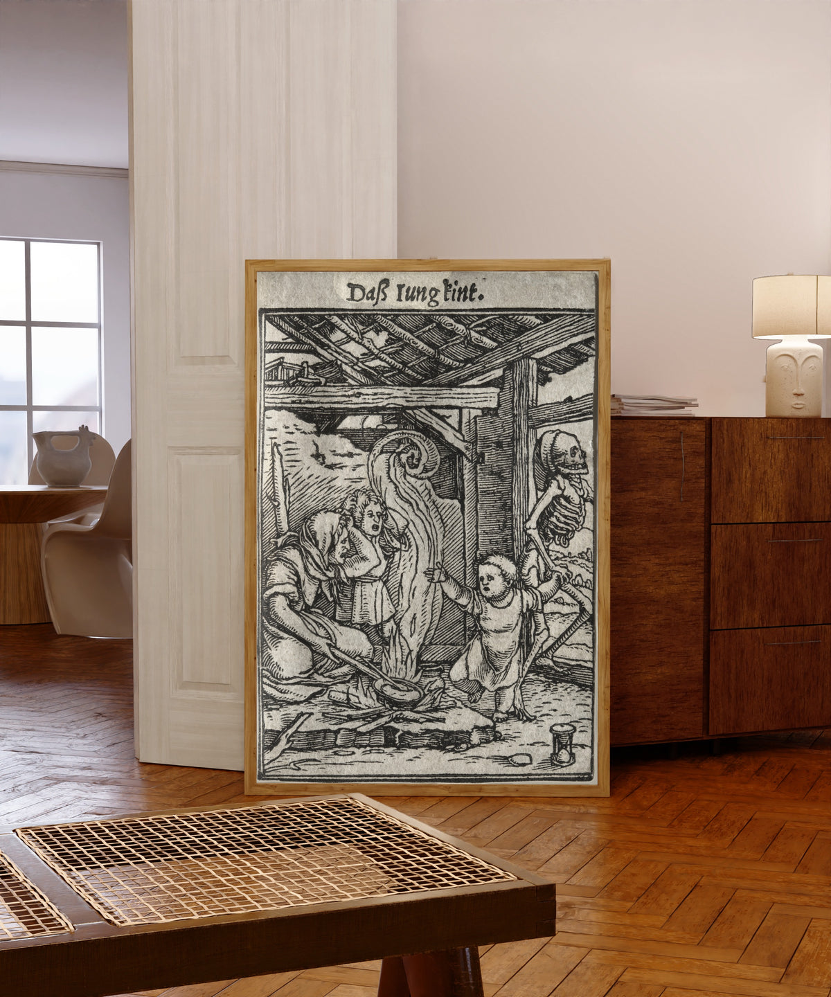 Framed art print of The Dance of Death: Daß Iung kint (The Child) by Hans Holbein the Younger, in a light wood frame leaning in a warm room with a parquet floor