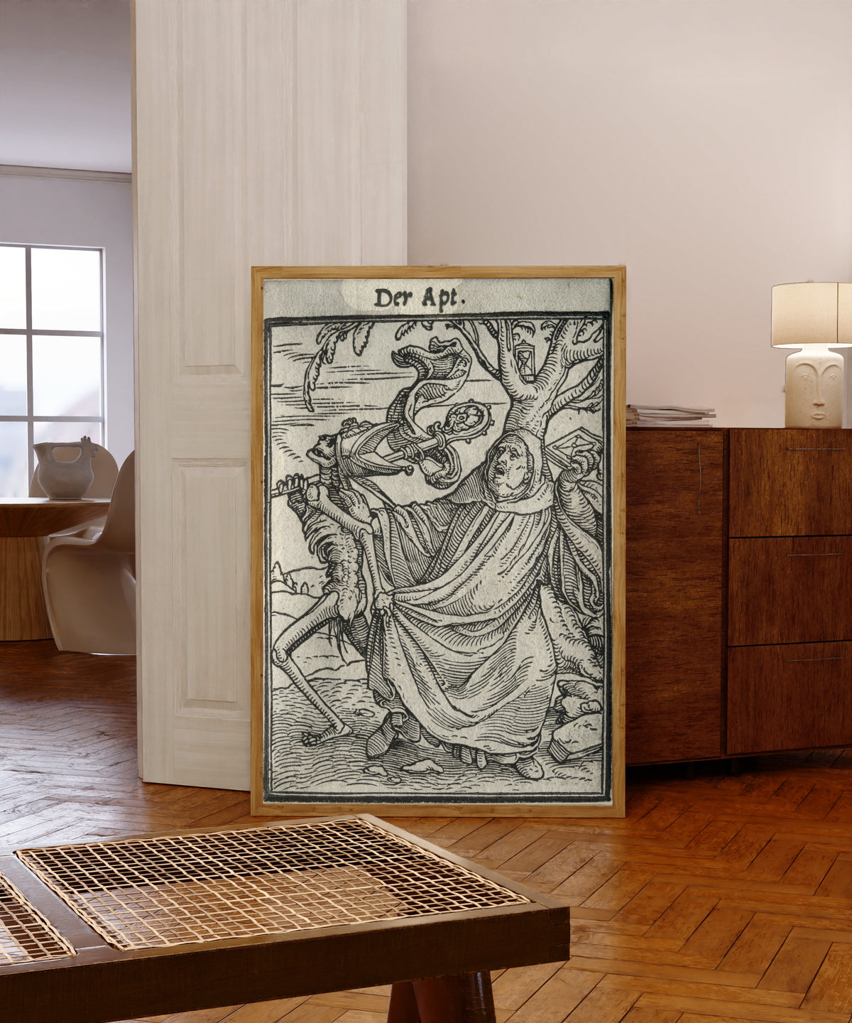 Framed art print of The Dance of Death: Der Apt (The Abbot) by Hans Holbein the Younger, in a light wood frame leaning in a warm room with a parquet floor