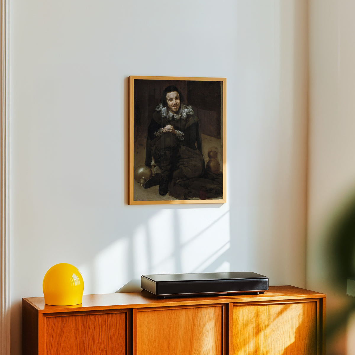 Framed art print of The Jester Calabacillas by Diego Velázquez, in a light wood frame on a white wall above an orange sideboard