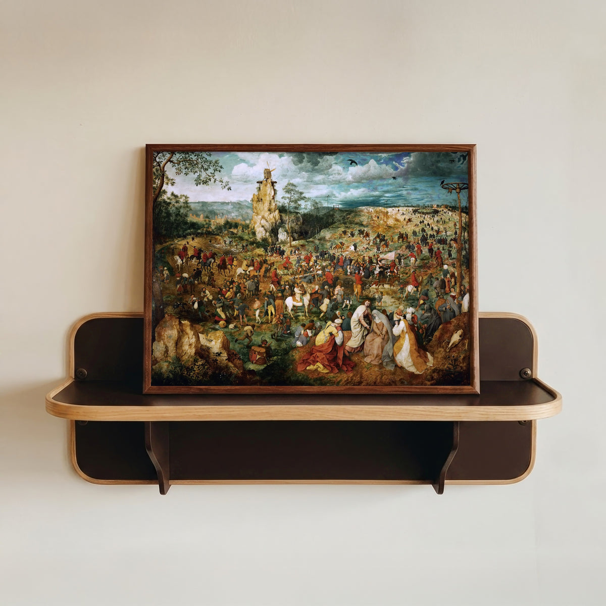 Framed art print of The Procession to Calvary by Pieter Bruegel the Elder, in a dark wood frame resting on a wall shelf