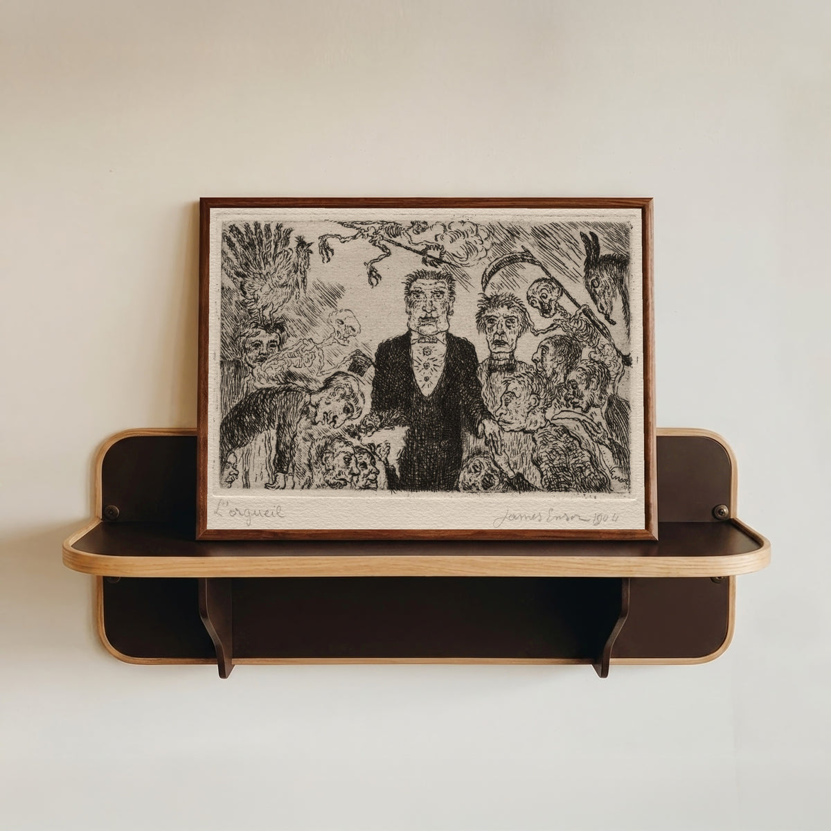 Framed art print of The Seven Deadly Sins: Pride by James Ensor, in a dark wood frame resting on a wall shelf