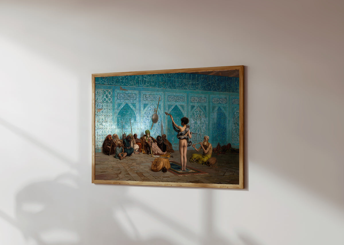Framed art print of The Snake Charmer by Jean-Léon Gérôme, in a light wood frame on a sunlit white wall, seen at a slight angle