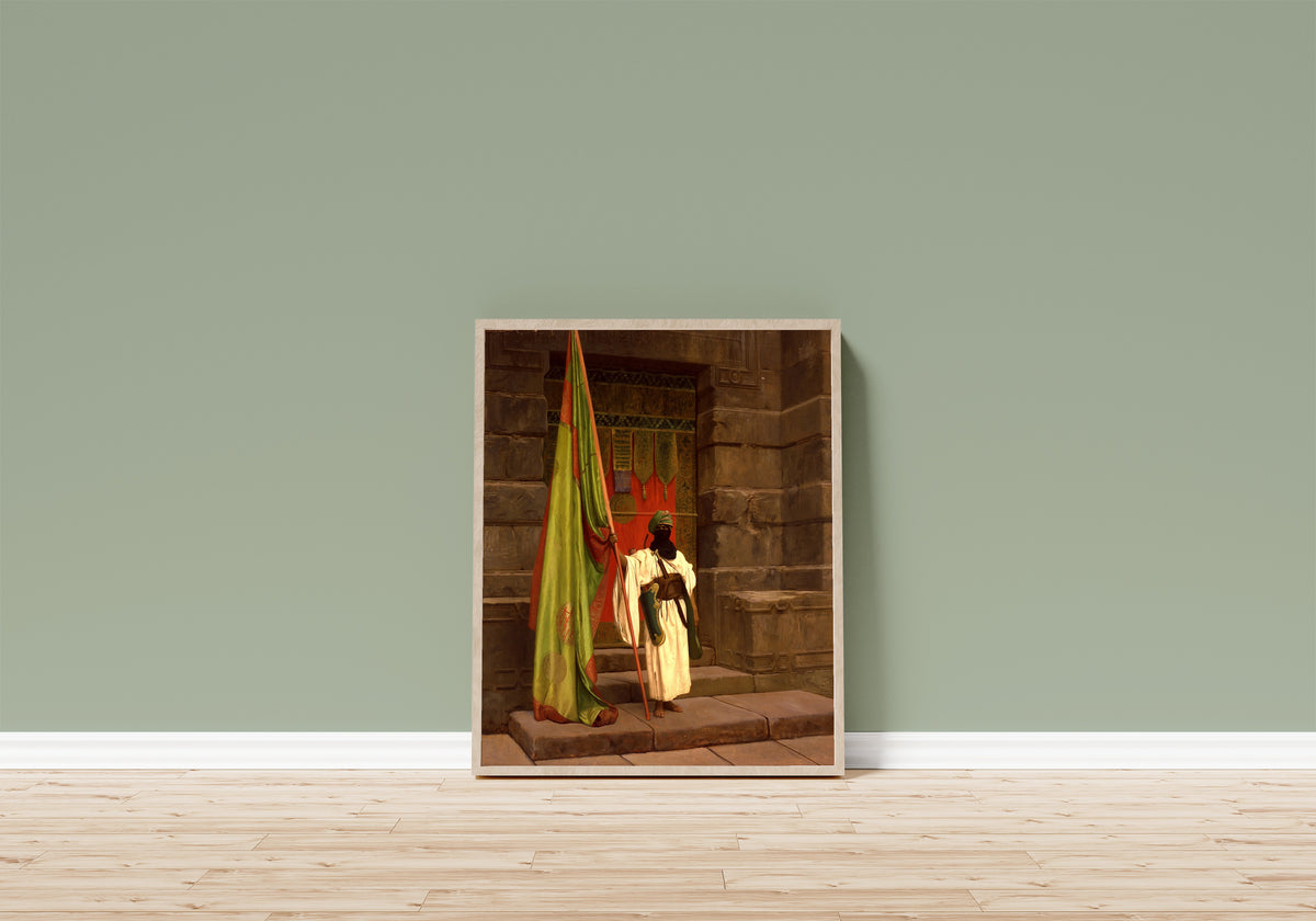 Framed art print of The Standing Bearer, Unfolding the Holy Flag by Jean-Léon Gérôme, in a light wood frame leaning on a sage green wall above a wooden floor
