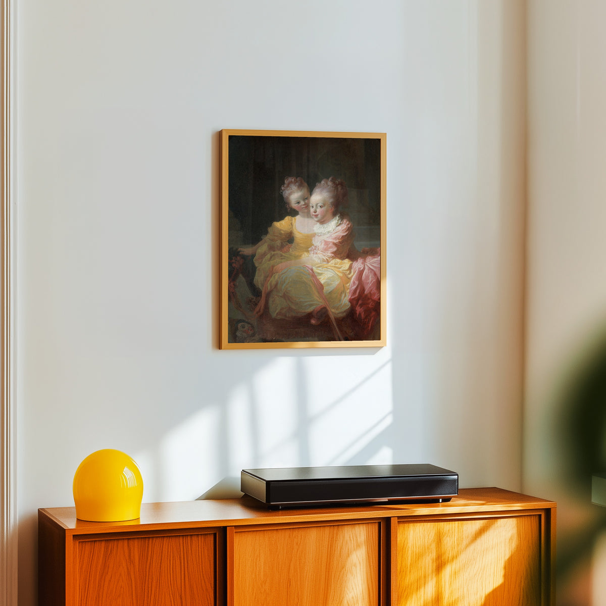 Framed art print of The Two Sisters by Jean-Honoré Fragonard, in a light wood frame on a white wall above an orange sideboard