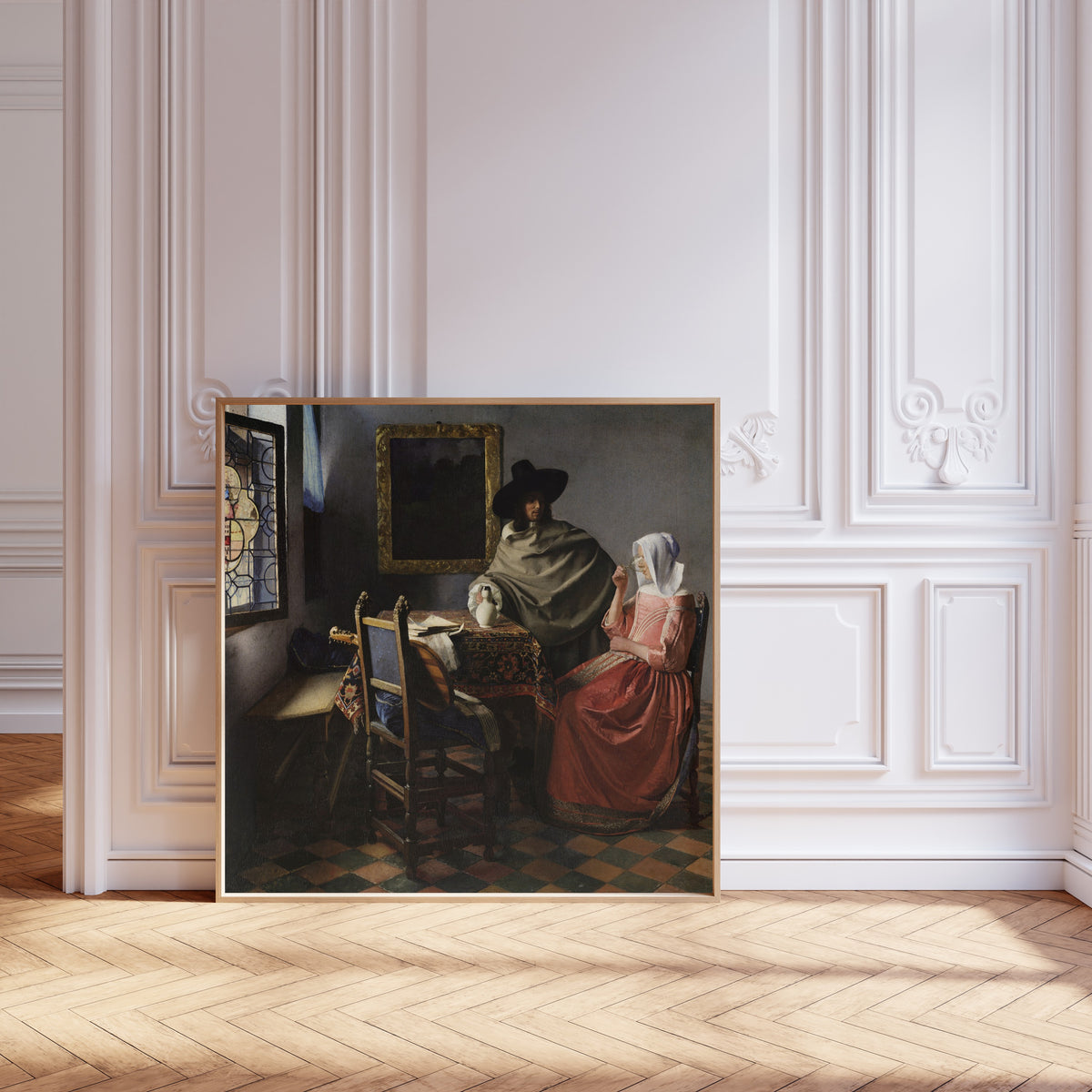 Framed art print of The Wine Glass by Johannes Vermeer, leaning against pale pink panelling on a herringbone floor
