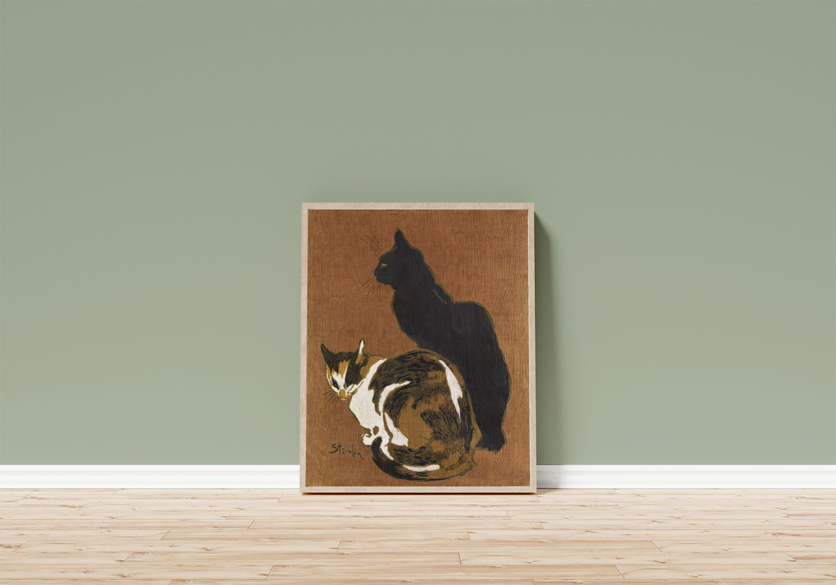 Framed art print of Two Cats by Théophile Steinlen, in a light wood frame leaning on a sage green wall above a wooden floor