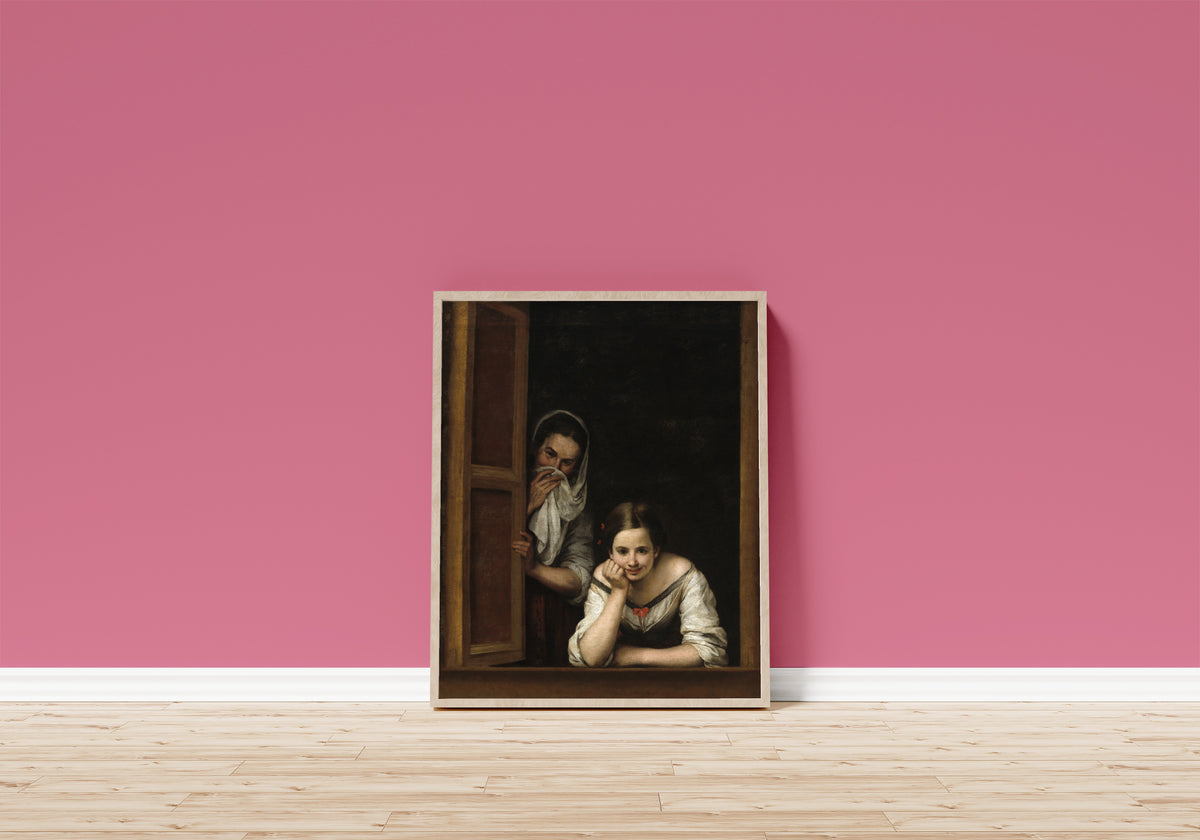 Framed art print of Two Women at a Window by Bartolomé Esteban Murillo, in a light wood frame leaning on a pink wall above a wooden floor