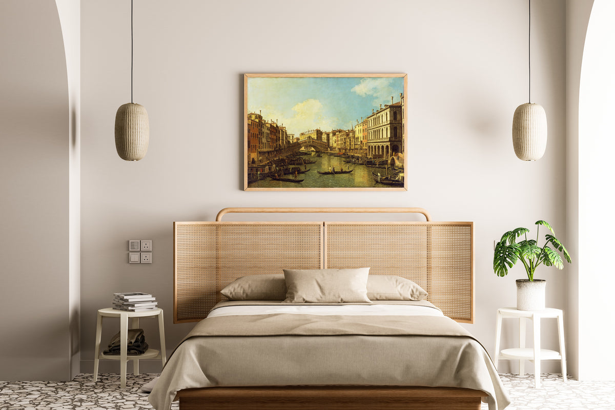 Framed art print of Venice, the Grand Canal from the Palazzo Dolfin-Manin to the Rialto Bridge by Canaletto, above a cane-headboard bed