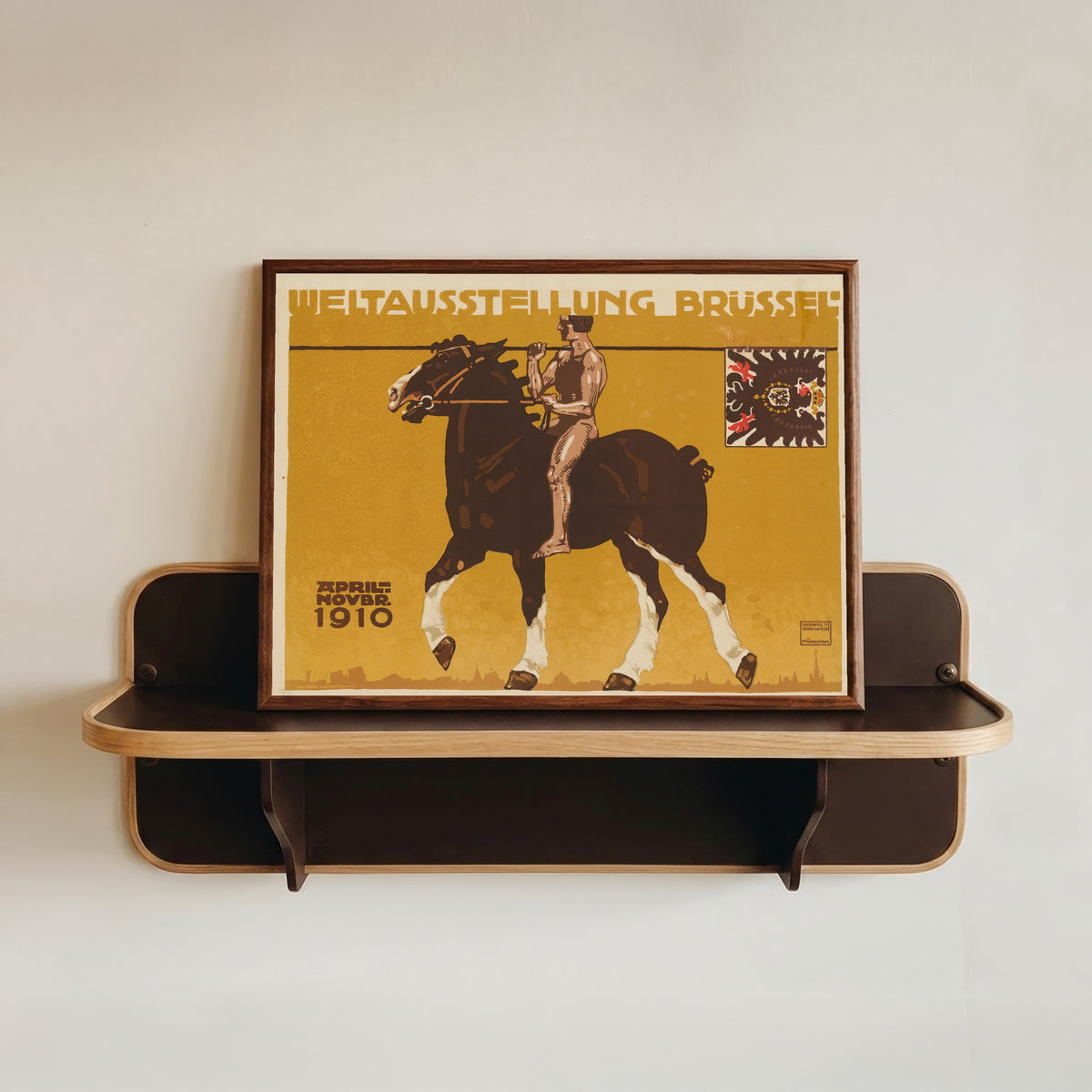 Framed art print of Weltausstellung Brüssel 1910 by Ludwig Hohlwein, in a dark wood frame resting on a wall shelf