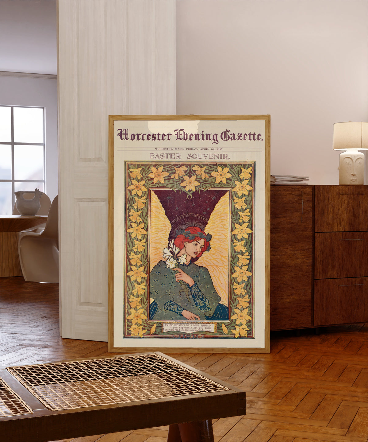 Framed art print of Worcester Evening Gazette, Easter Souvenir by Louis Rhead, in a light wood frame leaning in a warm room with a parquet floor
