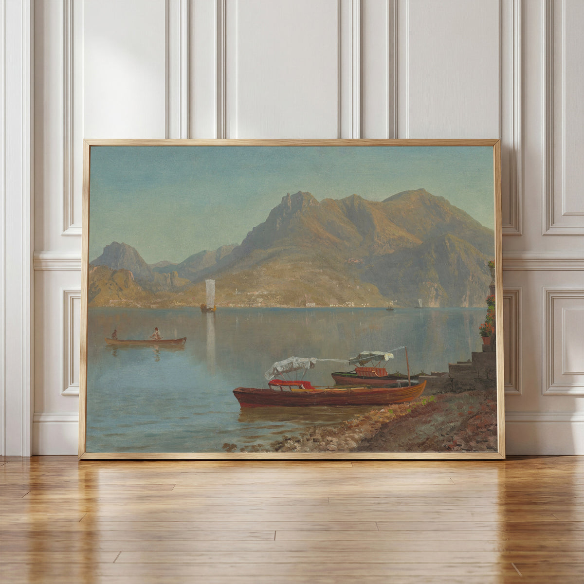 Framed art print of Lake Como by Albert Bierstadt, in a light wood frame leaning against a panelled white wall