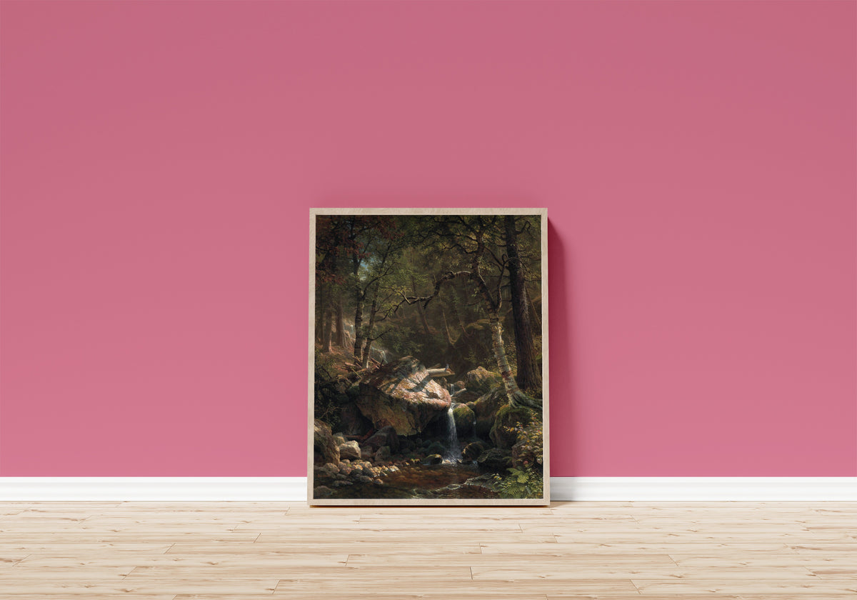 Framed art print of Mountain Brook by Albert Bierstadt, in a light wood frame leaning on a pink wall above a wooden floor