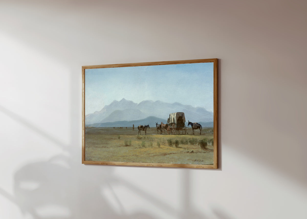 Framed art print of Surveyor's Wagon in the Rockies by Albert Bierstadt, in a light wood frame on a sunlit white wall, seen at a slight angle