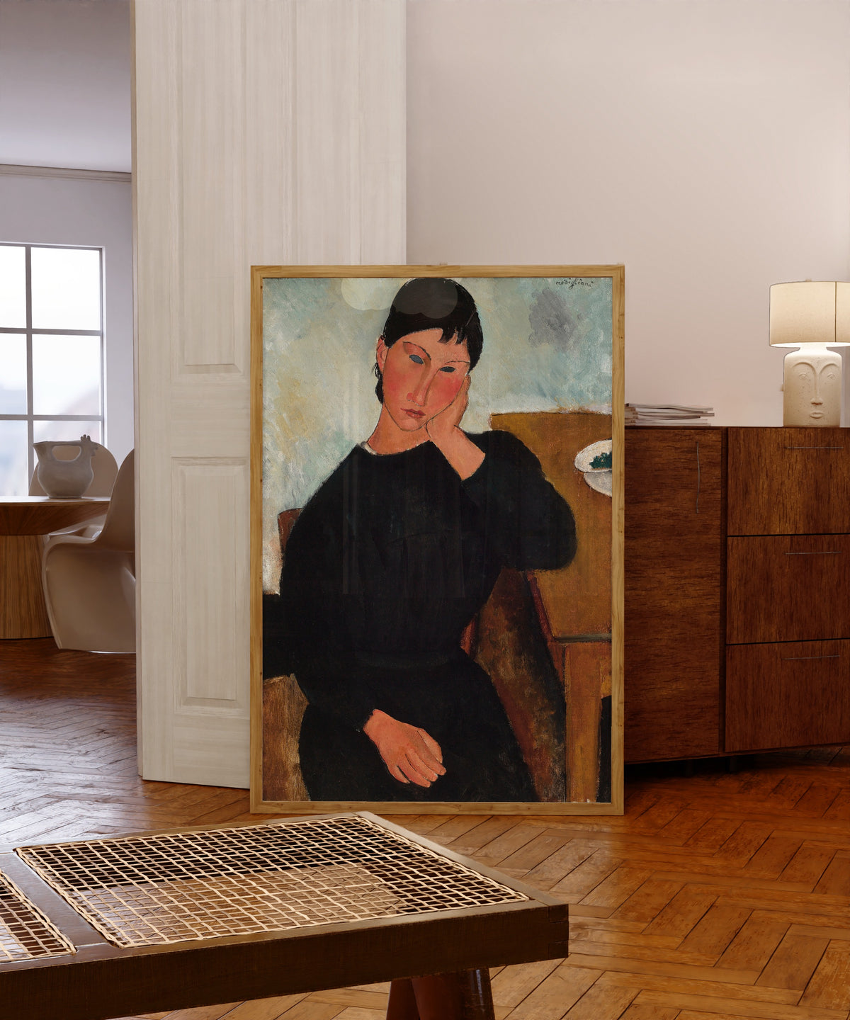 Framed art print of Elvira Resting at a Table by Amedeo Modigliani, in a light wood frame leaning in a warm room with a parquet floor