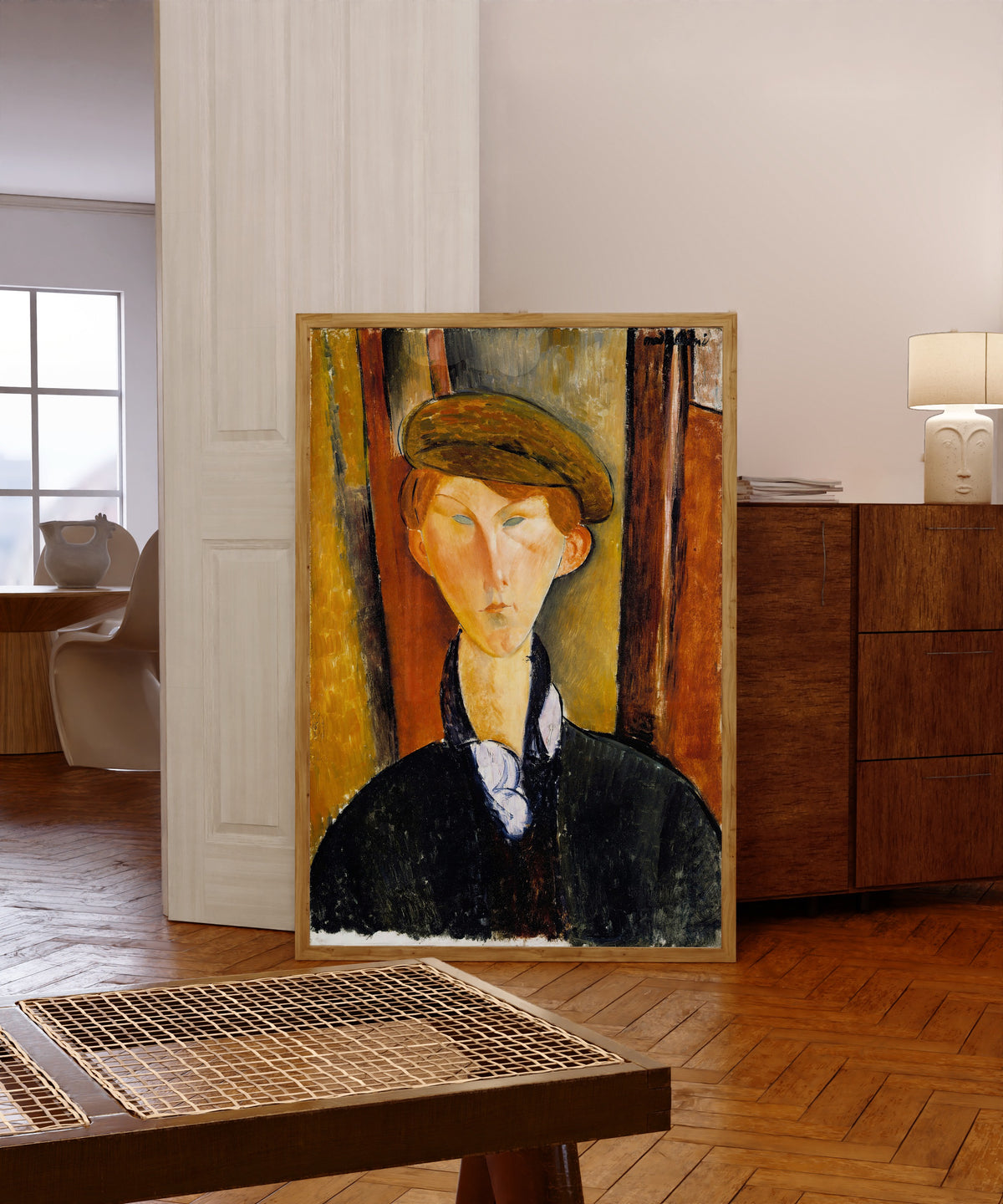 Framed art print of Young Man with a Cap by Amedeo Modigliani, in a light wood frame leaning in a warm room with a parquet floor