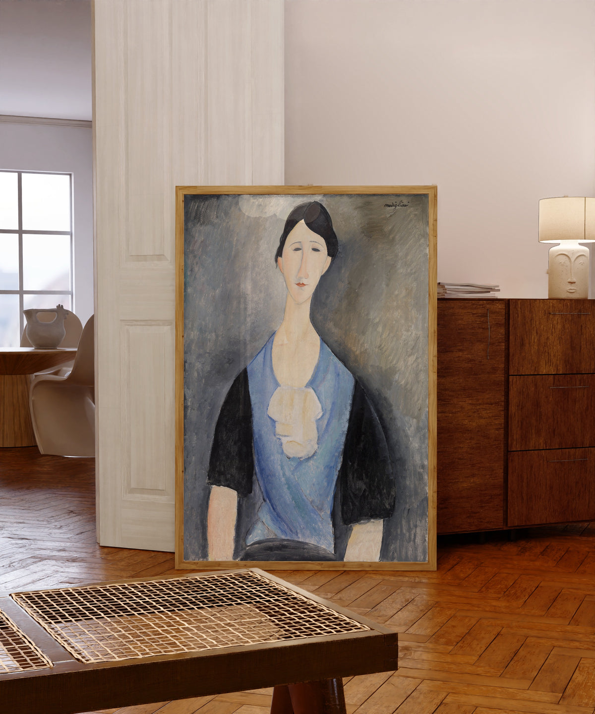 Framed art print of Young Woman in Blue by Amedeo Modigliani, in a light wood frame leaning in a warm room with a parquet floor