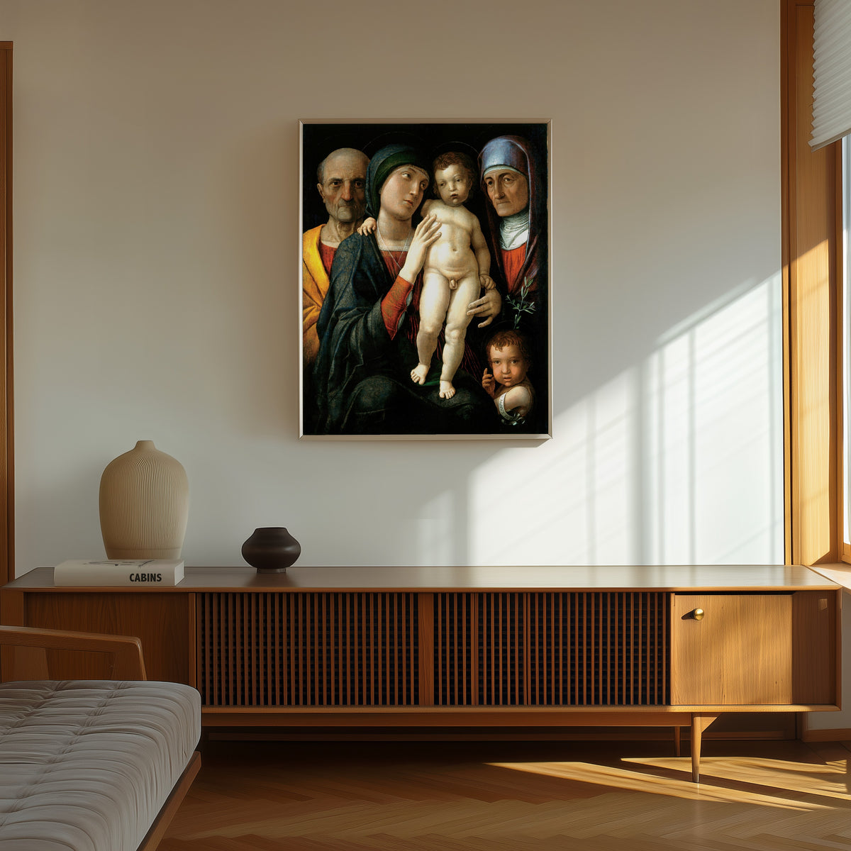 Framed art print of The Holy Family by Andrea Mantegna, in a thin light frame on a sunlit wall above a wooden sideboard