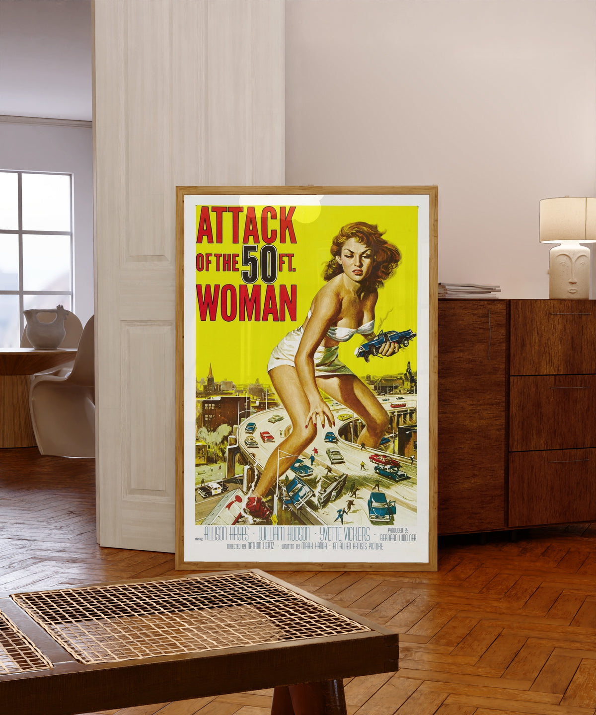 Framed art print of Attack of the 50 Foot Woman by Reynold Brown, in a light wood frame leaning in a warm room with a parquet floor