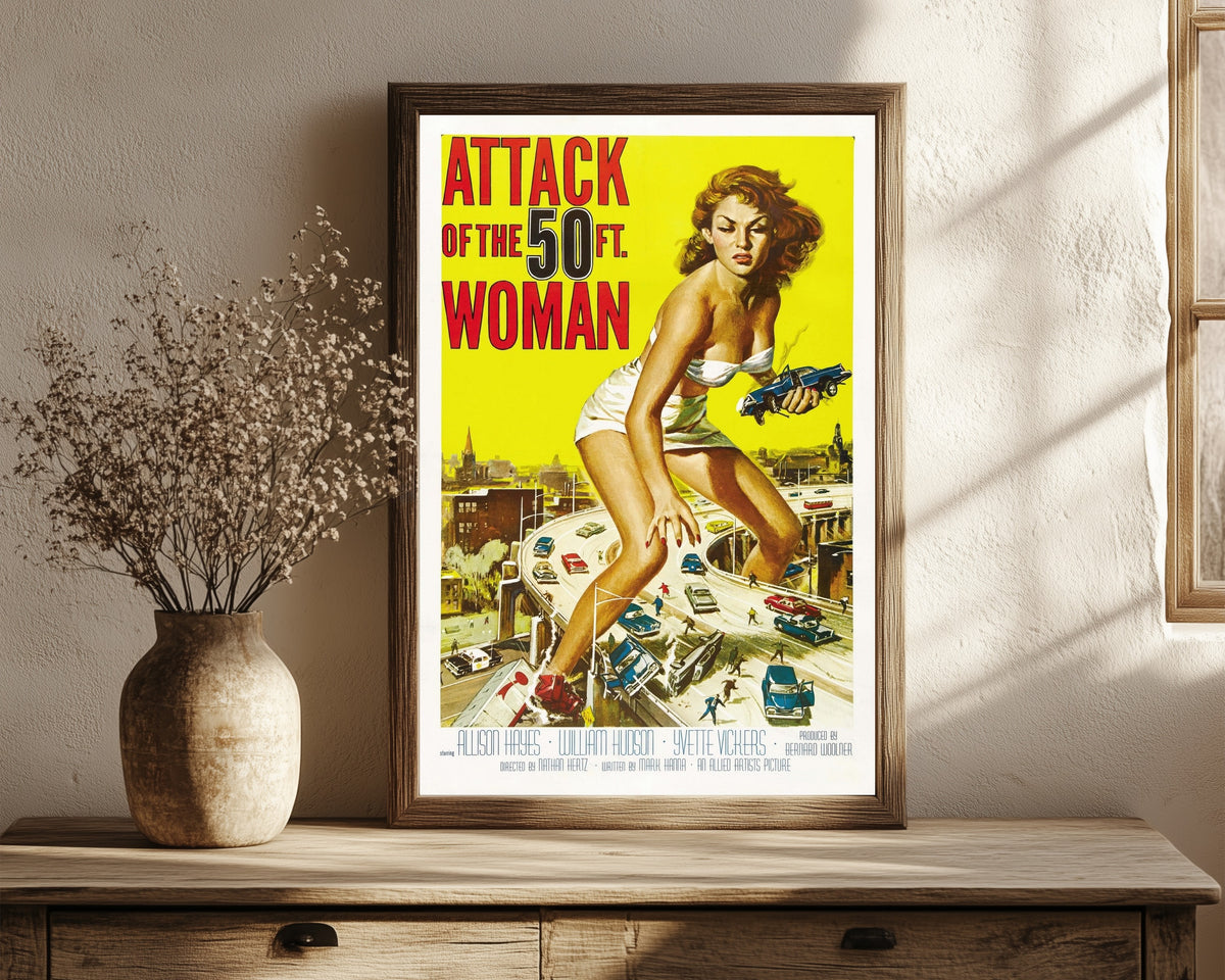 Framed art print of Attack of the 50 Foot Woman by Reynold Brown, in a dark wood frame leaning on a rustic console beside dried flowers