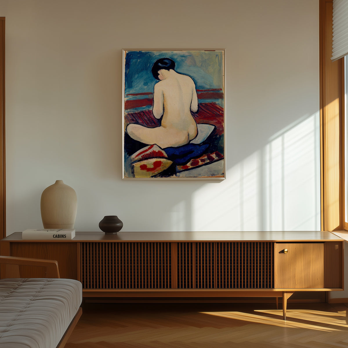 Framed art print of Sitting Nude with Pillow by August Macke, in a thin light frame on a sunlit wall above a wooden sideboard