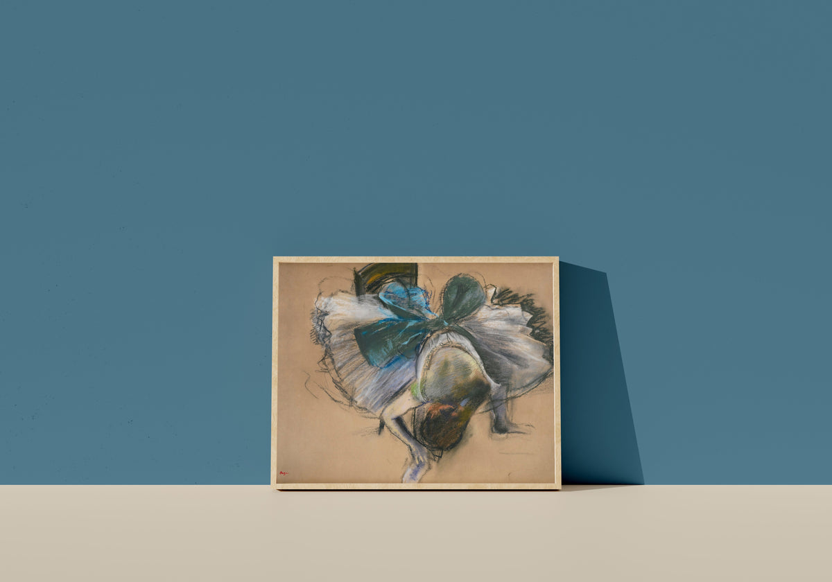 Framed art print of Dancer Adjusting Her Slipper (Blue and Green Bow) by Edgar Degas, in a light wood frame leaning on a blue wall
