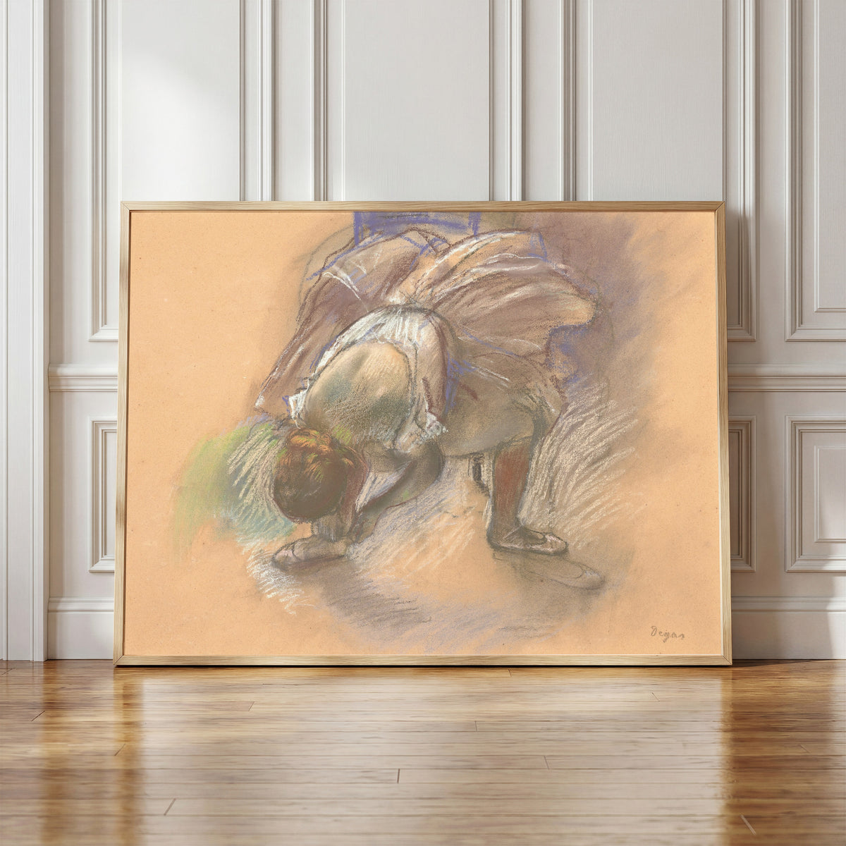 Framed art print of Dancer Adjusting Her Slipper (Lilac Tutu) by Edgar Degas, in a light wood frame leaning against a panelled white wall