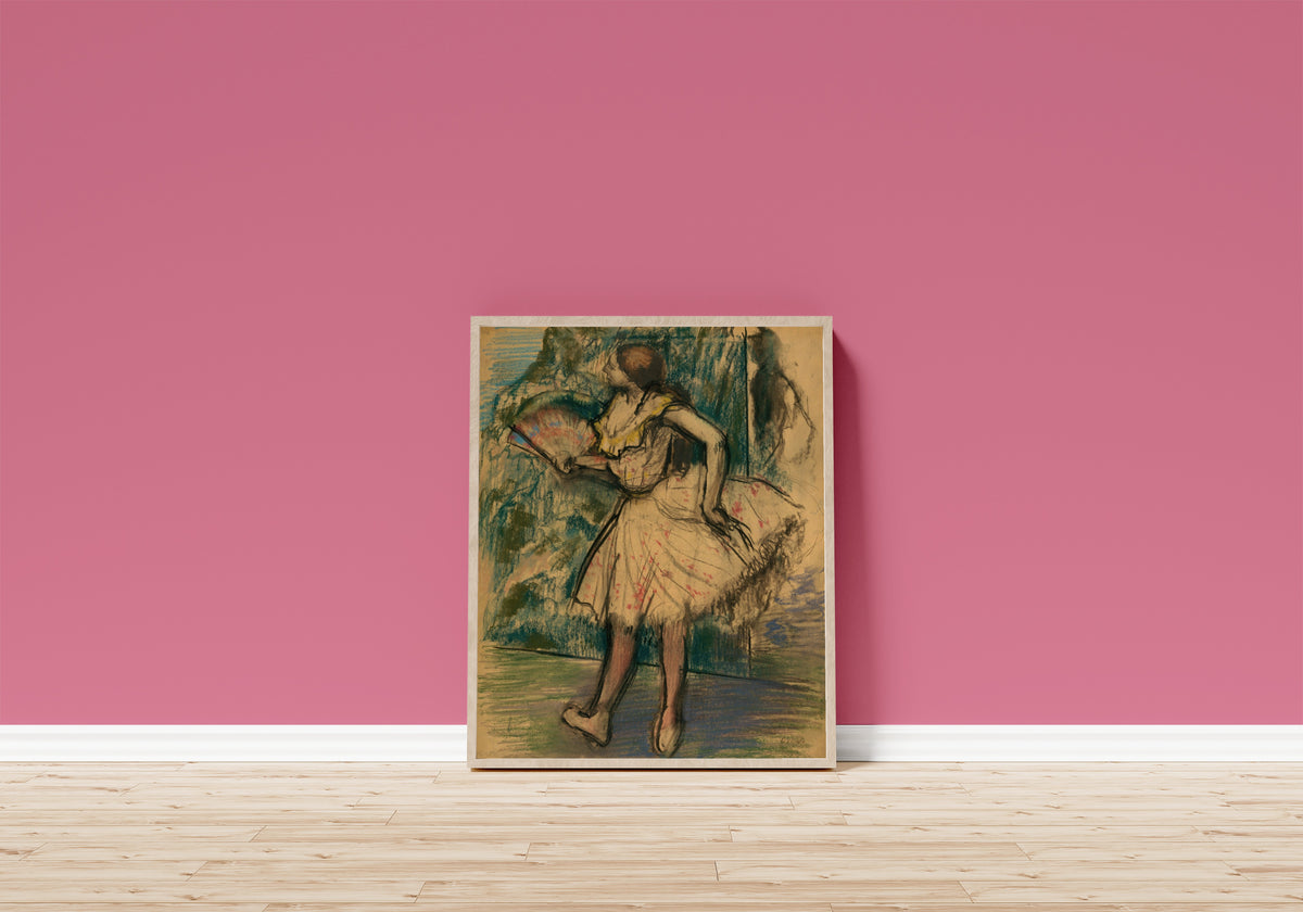 Framed art print of Dancer with a Fan by Edgar Degas, in a light wood frame leaning on a pink wall above a wooden floor