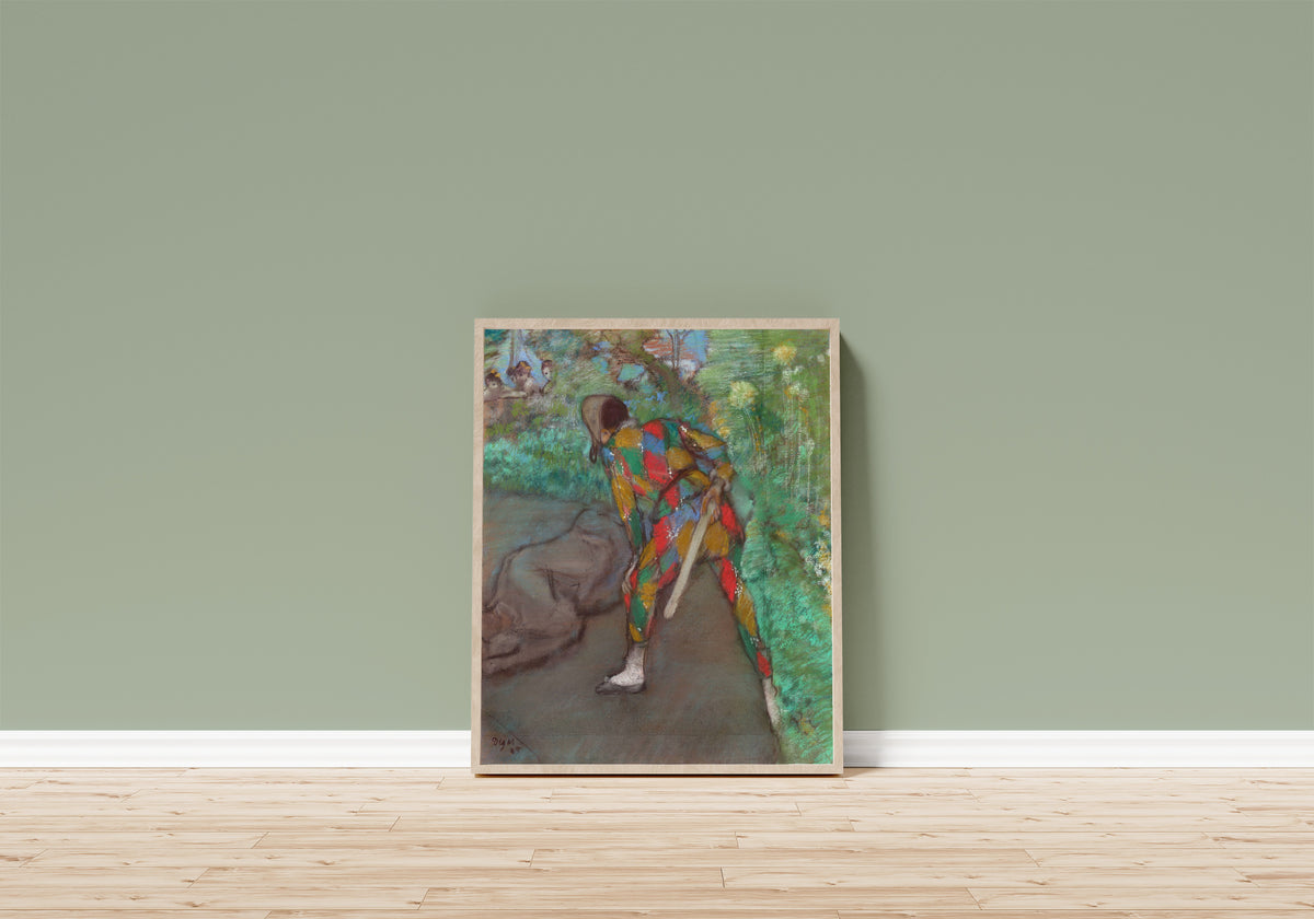 Framed art print of Harlequin by Edgar Degas, in a light wood frame leaning on a sage green wall above a wooden floor