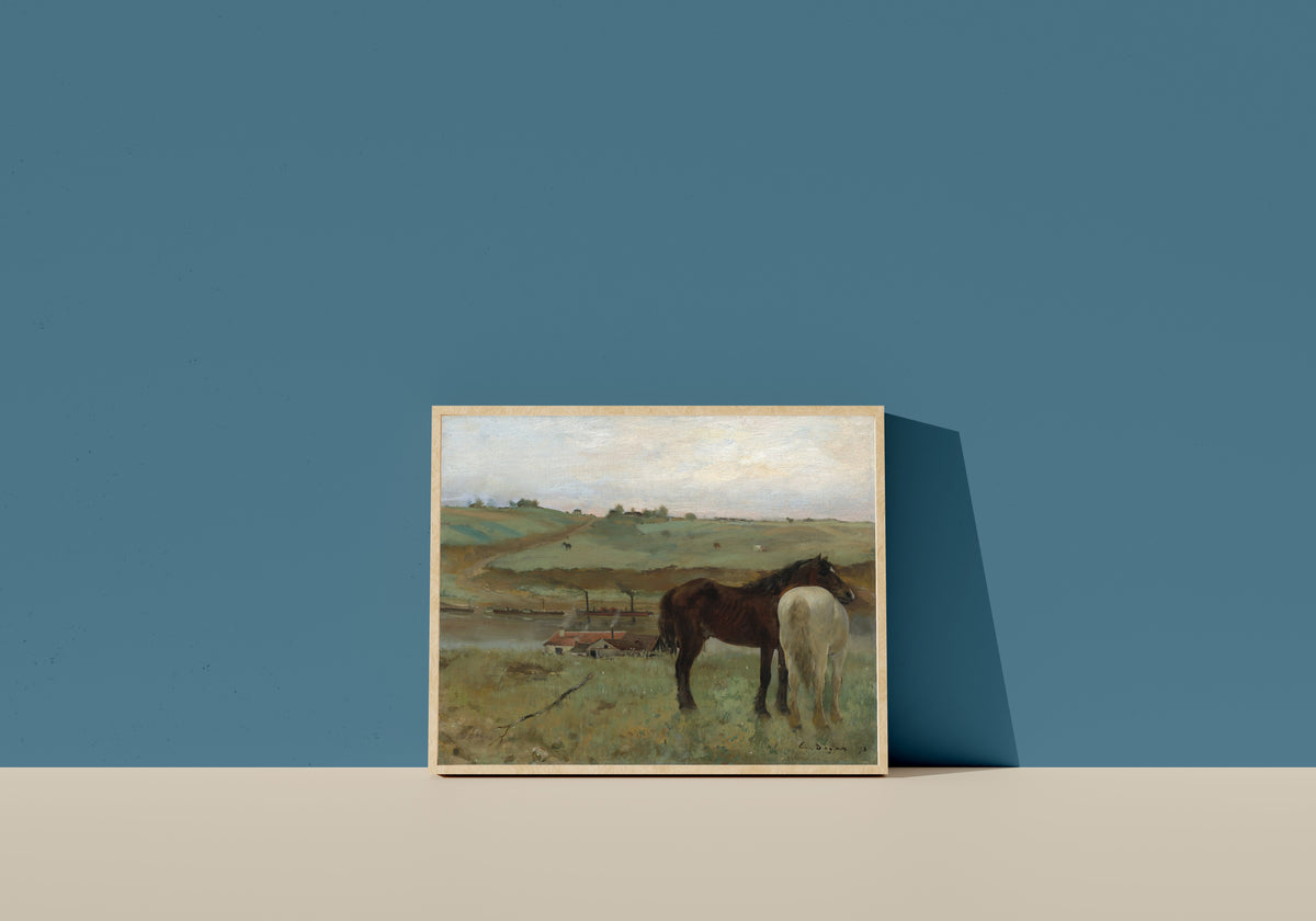 Framed art print of Horses in a Meadow by Edgar Degas, in a light wood frame leaning on a blue wall