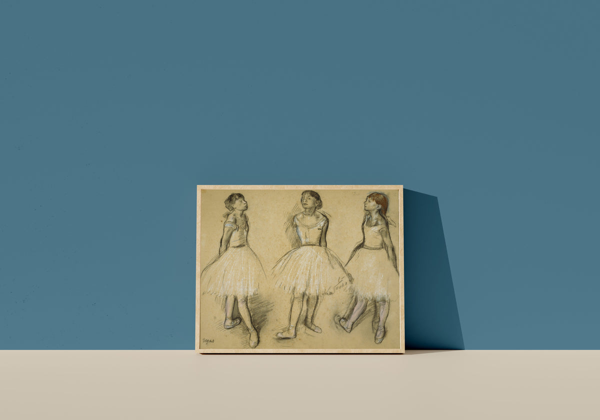Framed art print of Three Studies of a Dancer in Fourth Position by Edgar Degas, in a light wood frame leaning on a blue wall