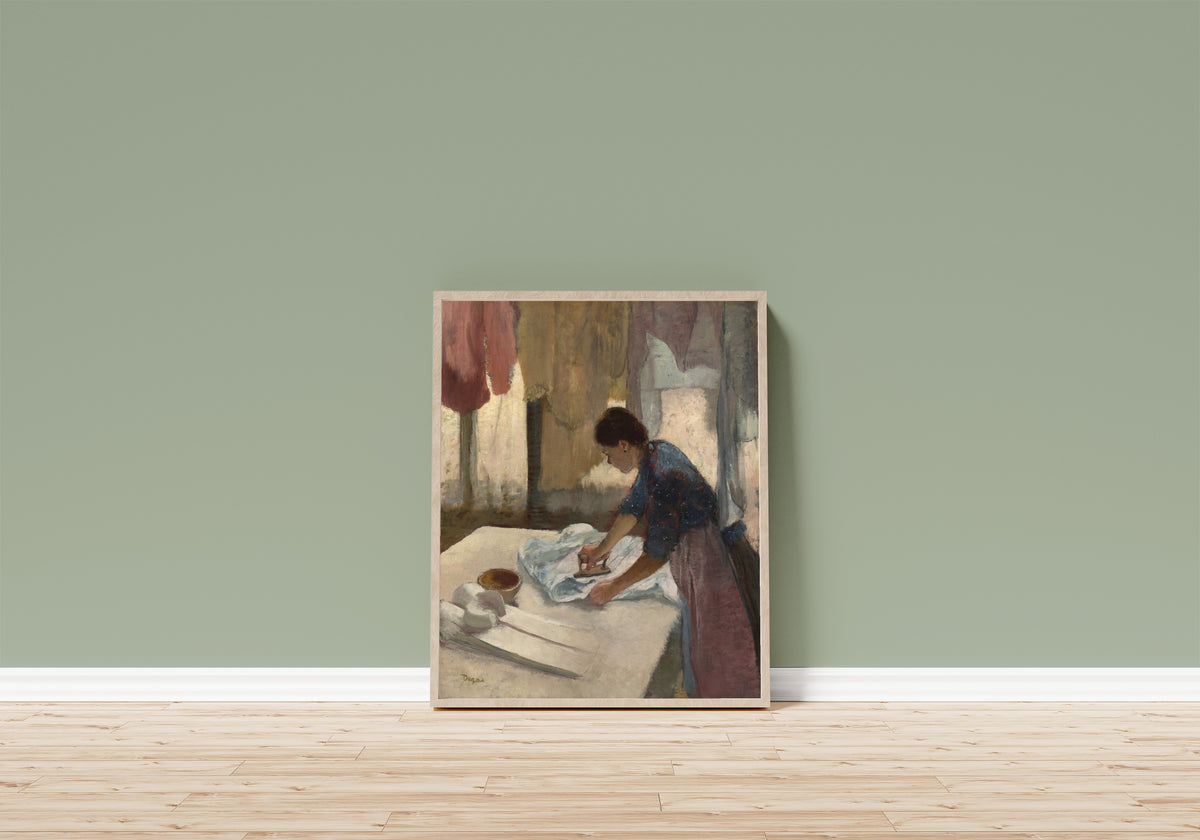 Framed art print of Woman Ironing in a Blue Blouse by Edgar Degas, in a light wood frame leaning on a sage green wall above a wooden floor