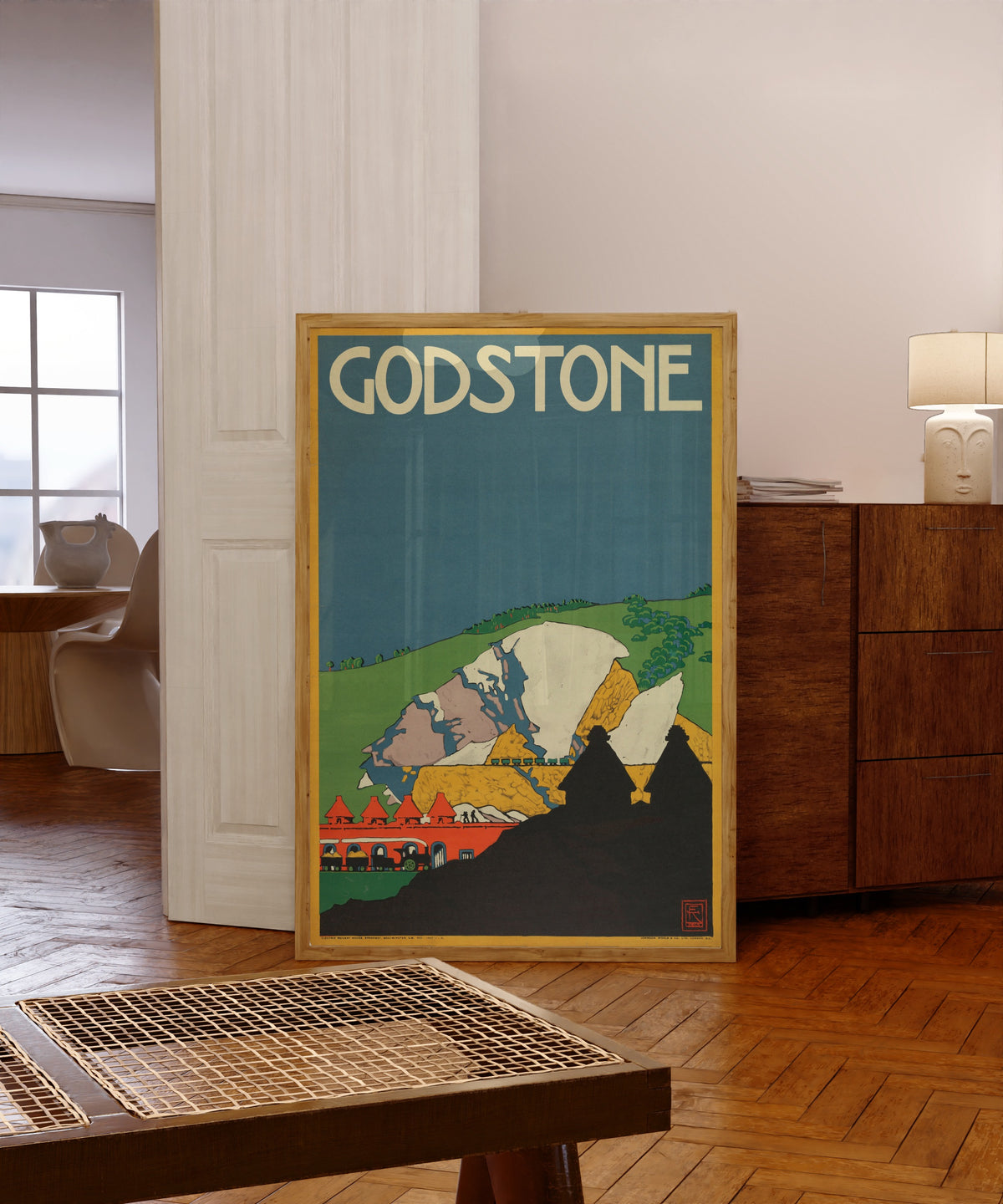 Framed art print of Godstone by Edward McKnight Kauffer, in a light wood frame leaning in a warm room with a parquet floor
