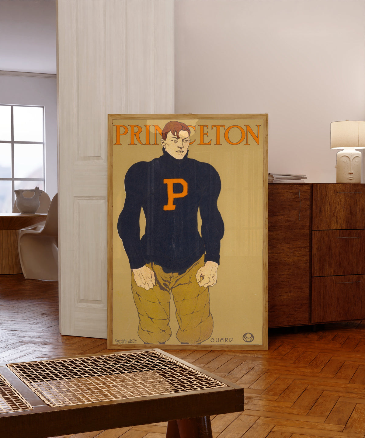 Framed art print of Princeton by Edward Penfield, in a light wood frame leaning in a warm room with a parquet floor