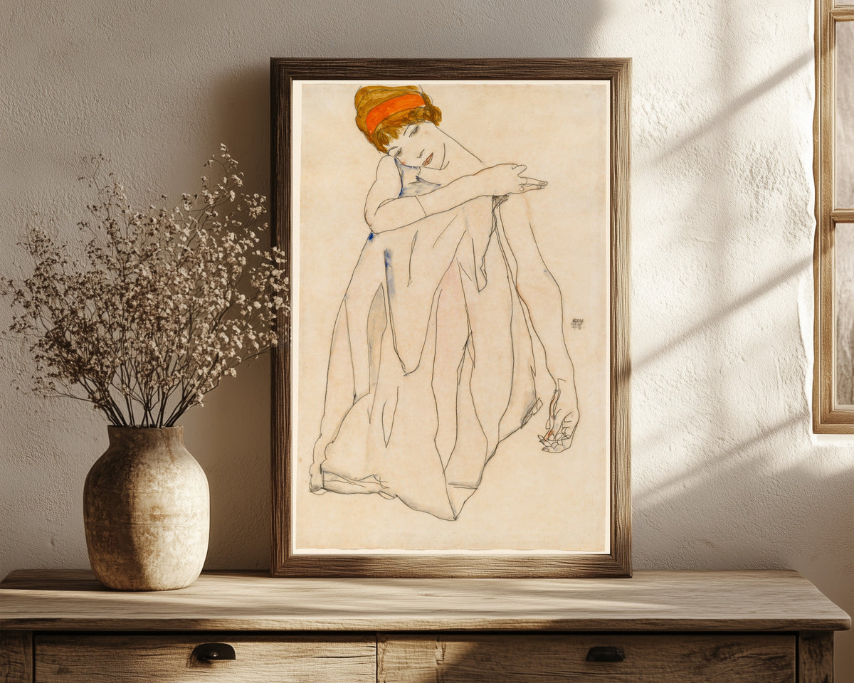 Framed art print of Dancer (Die Tänzerin) by Egon Schiele, in a dark wood frame leaning on a rustic console beside dried flowers