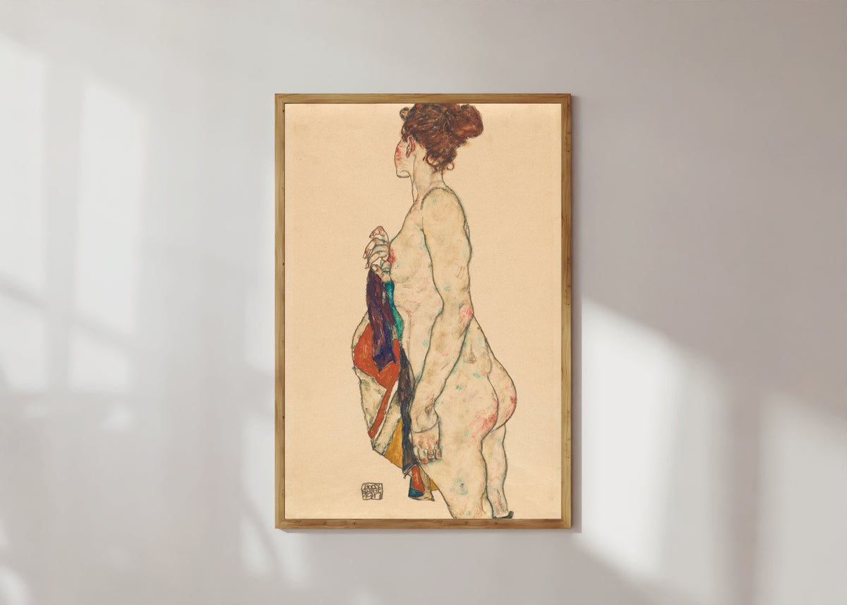 Framed art print of Standing Nude with a Patterned Robe by Egon Schiele, in a light wood frame on a sunlit white wall