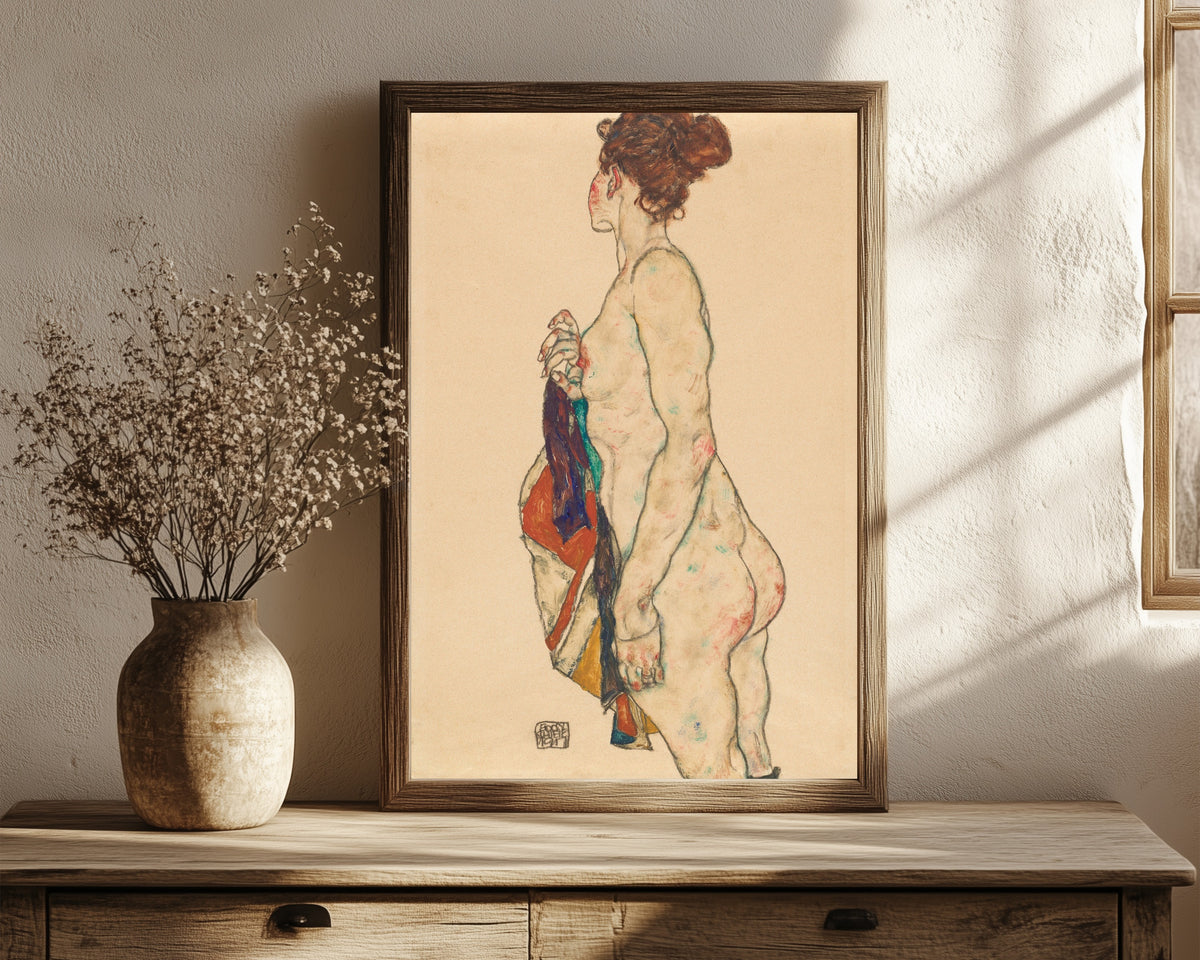 Framed art print of Standing Nude with a Patterned Robe by Egon Schiele, in a dark wood frame leaning on a rustic console beside dried flowers