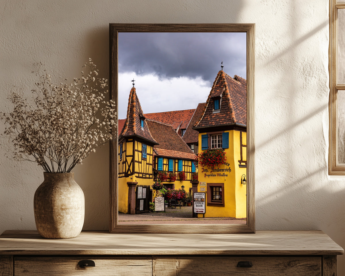 Framed art print of Eguisheim (Alsace) by Daniela Faust, in a dark wood frame leaning on a rustic console beside dried flowers