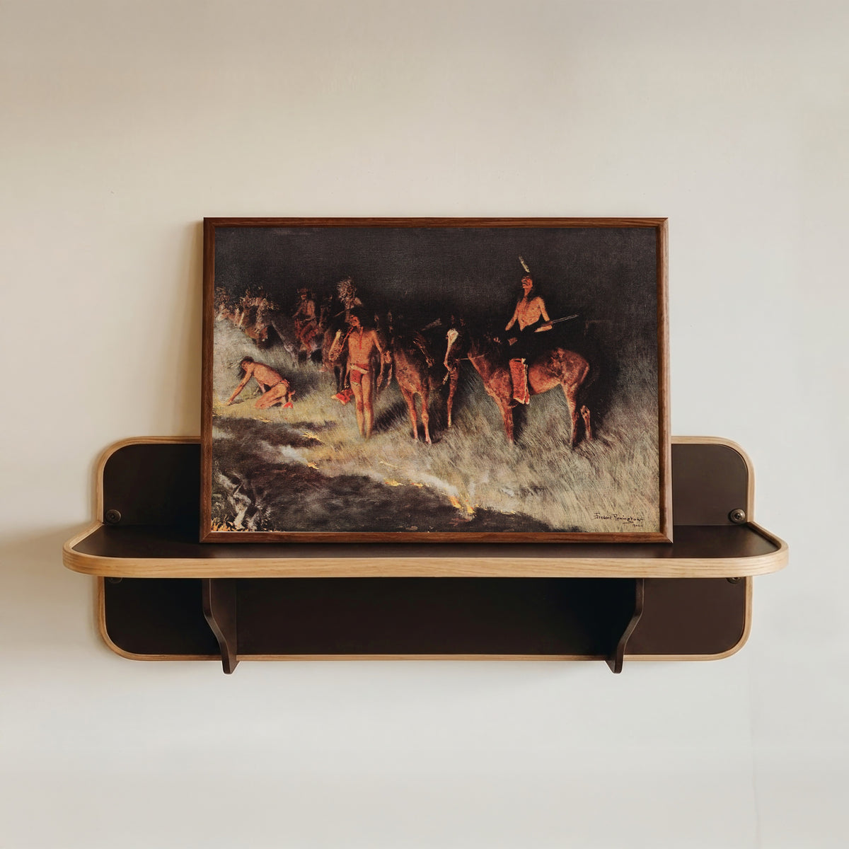 Framed art print of The Grass Fire by Frederic Remington, in a dark wood frame resting on a wall shelf