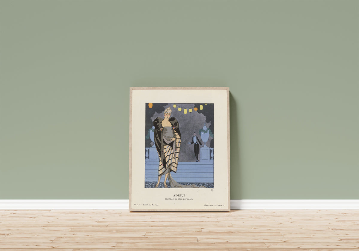 Framed art print of Adieu! Manteau du soir de Worth by George Barbier, in a light wood frame leaning on a sage green wall above a wooden floor