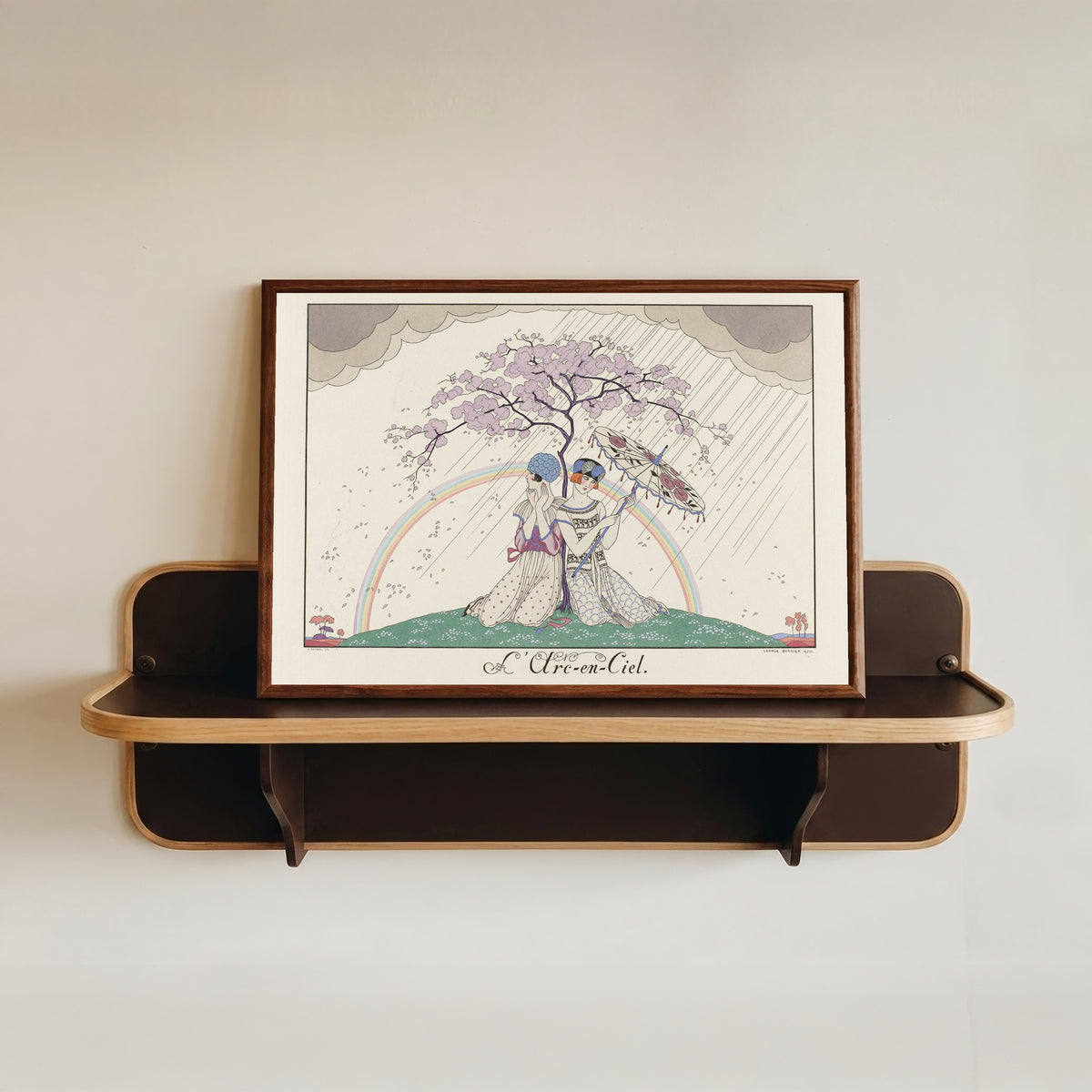 Framed art print of L'Arc-en-ciel [The Rainbow] by George Barbier, in a dark wood frame resting on a wall shelf