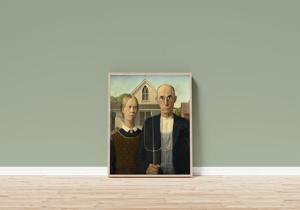 Framed art print of American Gothic by Grant Wood, in a light wood frame leaning on a sage green wall above a wooden floor